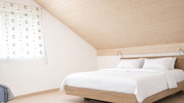 A bedroom with a sloped ceiling, a wooden bed frame, white bedding, and a window with a geometric pattern. - Delightful 1-bedroom apartment in Safenwil perfect for your stay (Safenwil)