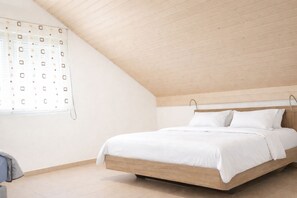 Une chambre avec un plafond en pente, un cadre de lit en bois, du linge de lit blanc et une fenêtre ornée d’un motif géométrique.