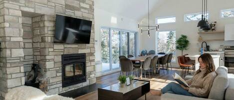 A modern living room with a stone fireplace, a flat-screen TV, a dining area, and a kitchen with pendant lighting.