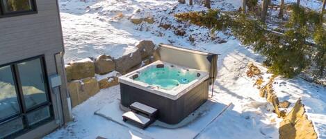 Un bain à remous installé dans un décor extérieur enneigé, avec un mur en pierre et un bâtiment doté de grandes fenêtres.