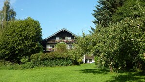 A two-story house with a balcony, surrounded by lush greenery and trees.