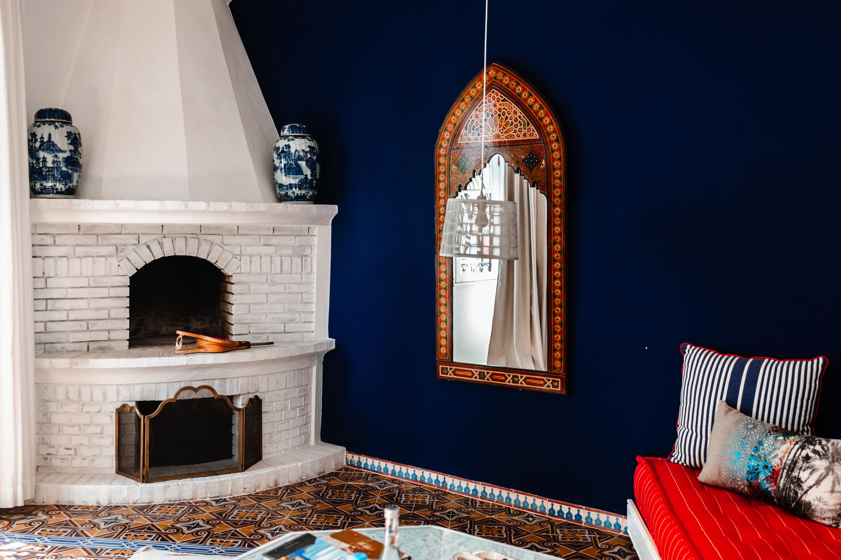 A room with a white brick fireplace, blue and white patterned tiles, a red sofa, and a large ornate mirror.