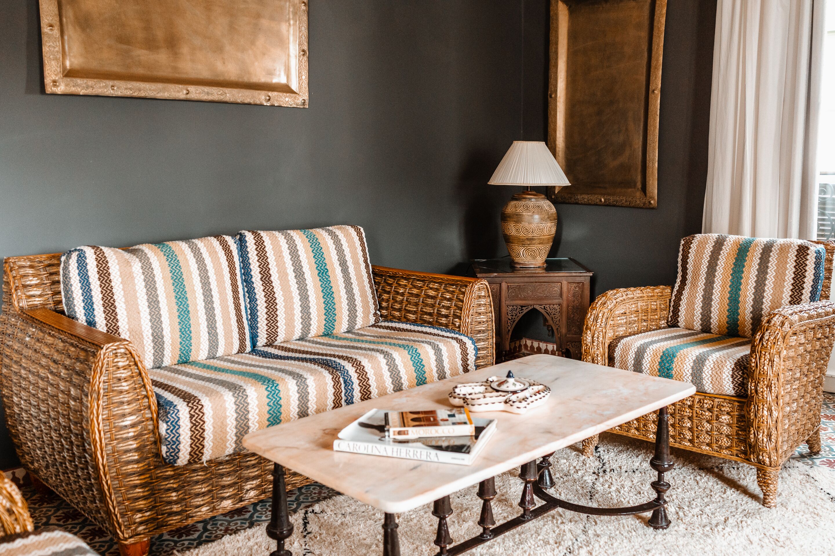 A room with a wicker sofa, a matching armchair, a small wooden side table with a lamp, and a marble coffee table.