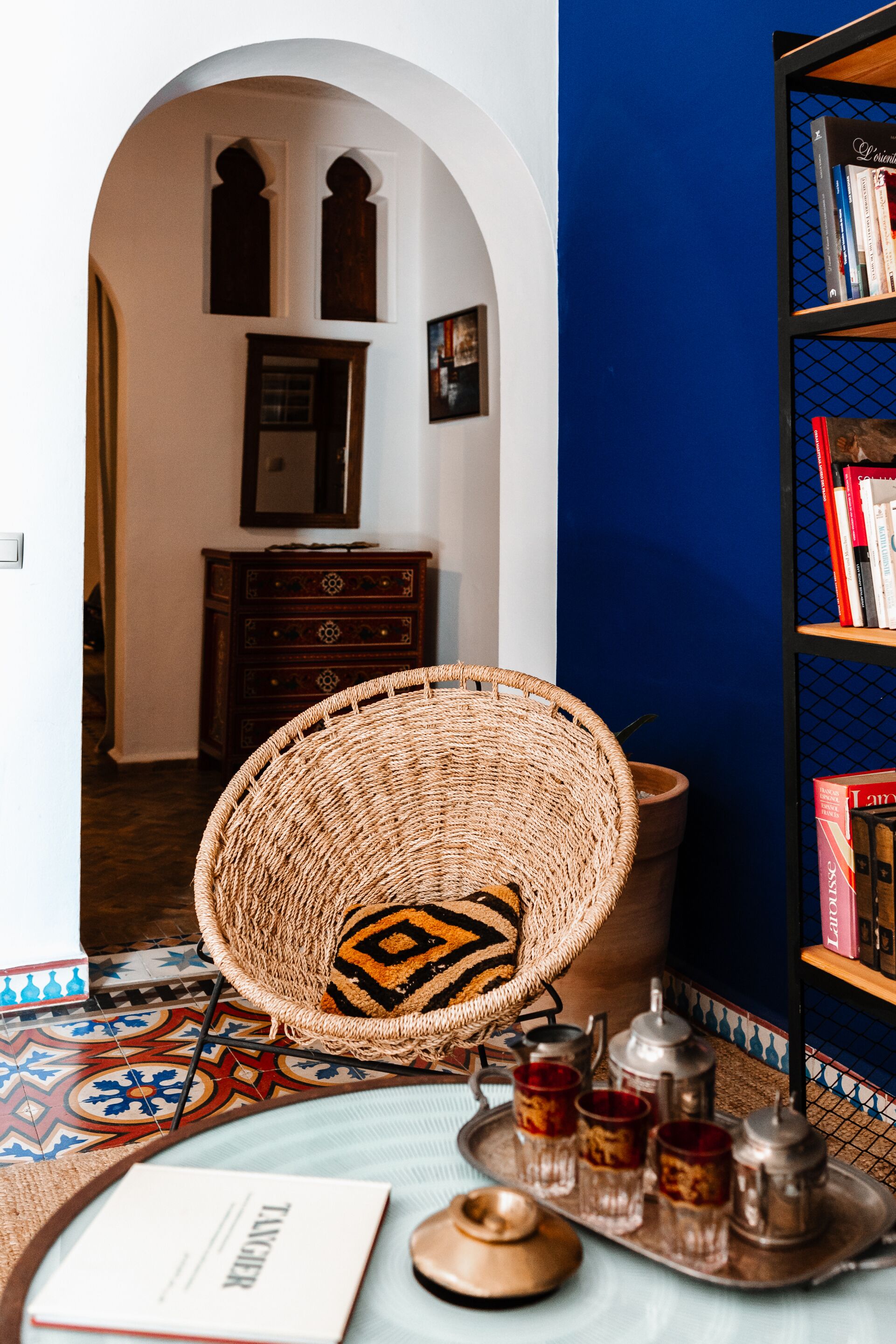 A room with a woven chair, a bookshelf, a tray with teapots and cups, and a patterned rug.