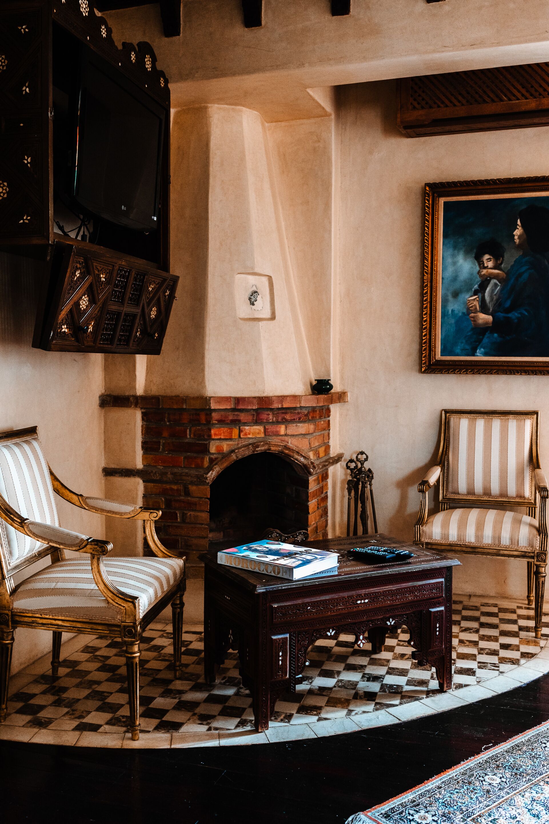 A room with a checkered floor, a fireplace, two upholstered chairs, a coffee table, a television, and a framed painting on the wall.