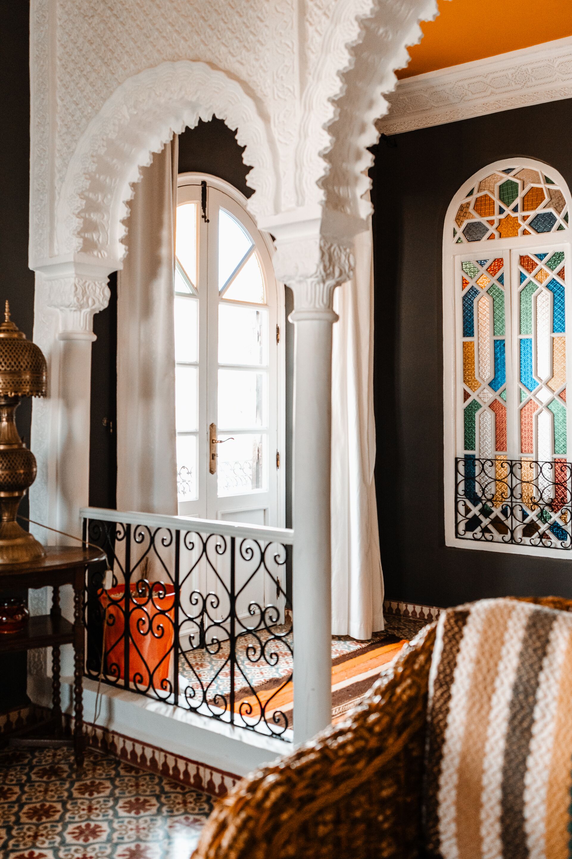 Une pièce dotée d’un sol à motifs, d’une porte en arc blanc, d’une fenêtre ornée de vitraux colorés et d’une rambarde en métal.