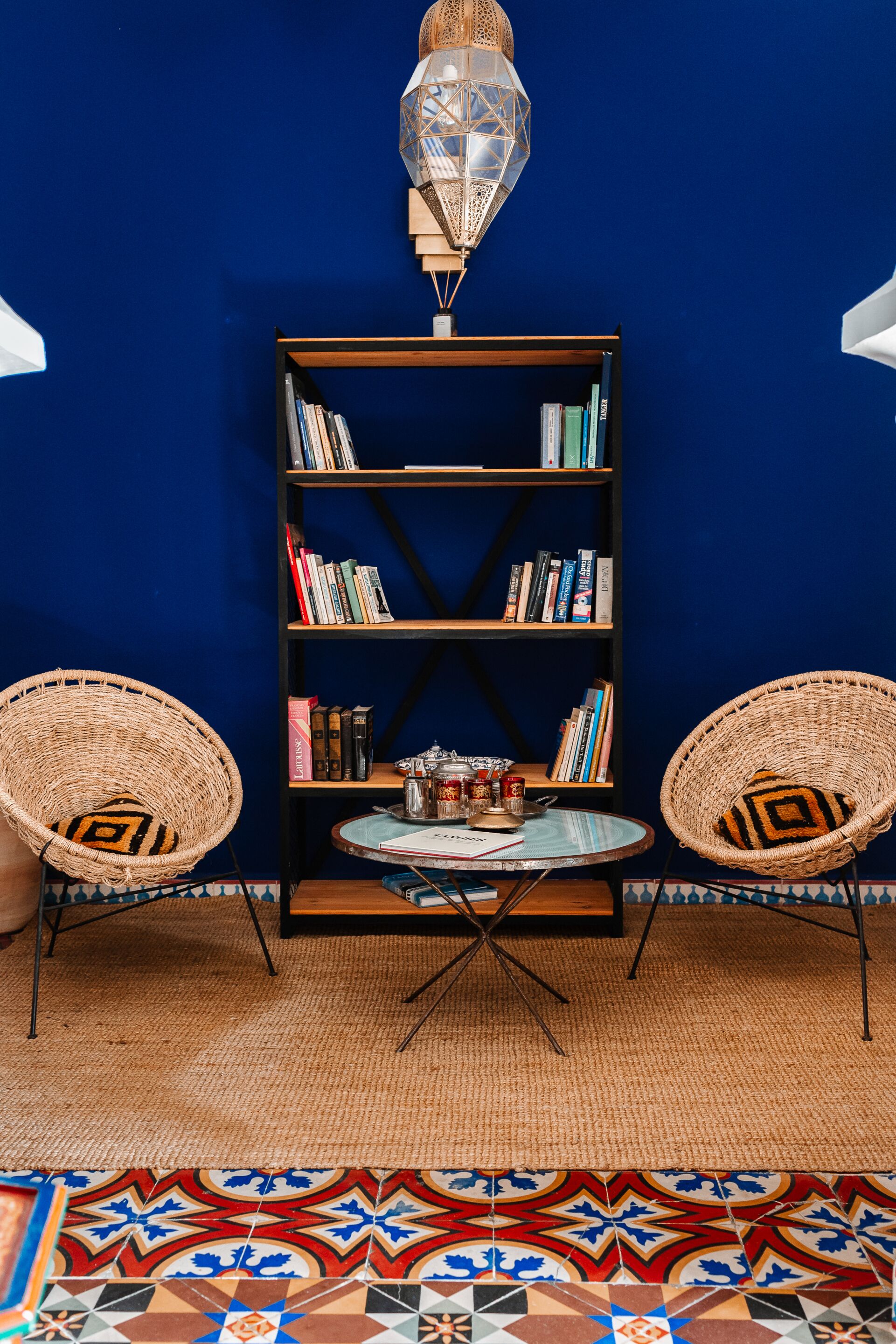 A room with a blue wall, a bookshelf, two wicker chairs, a small round table, and a patterned rug.