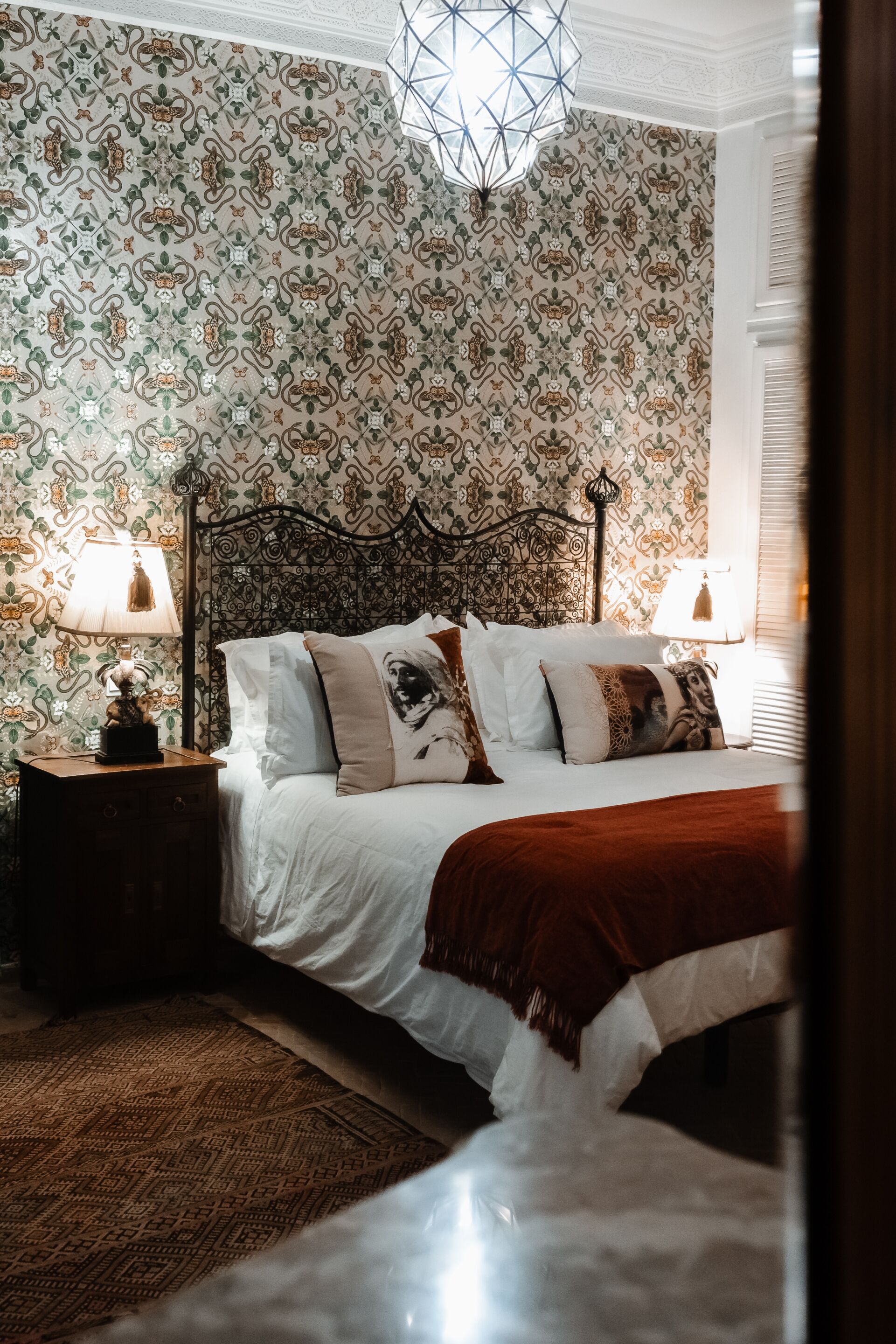 A bedroom with a patterned wallpaper, a bed with multiple pillows, a nightstand with a lamp, and a geometric ceiling light.