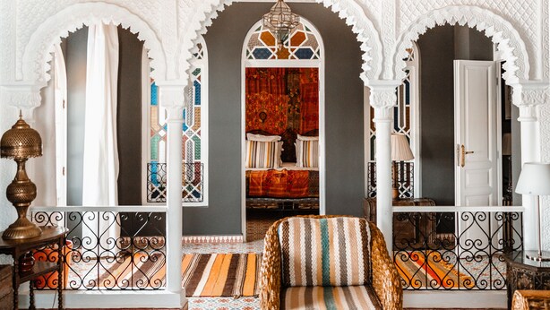 A room with a patterned sofa, a decorative lamp, and a view through an arched doorway to a bed with a red and orange bedspread. - Lalla Soulika (Tangier)