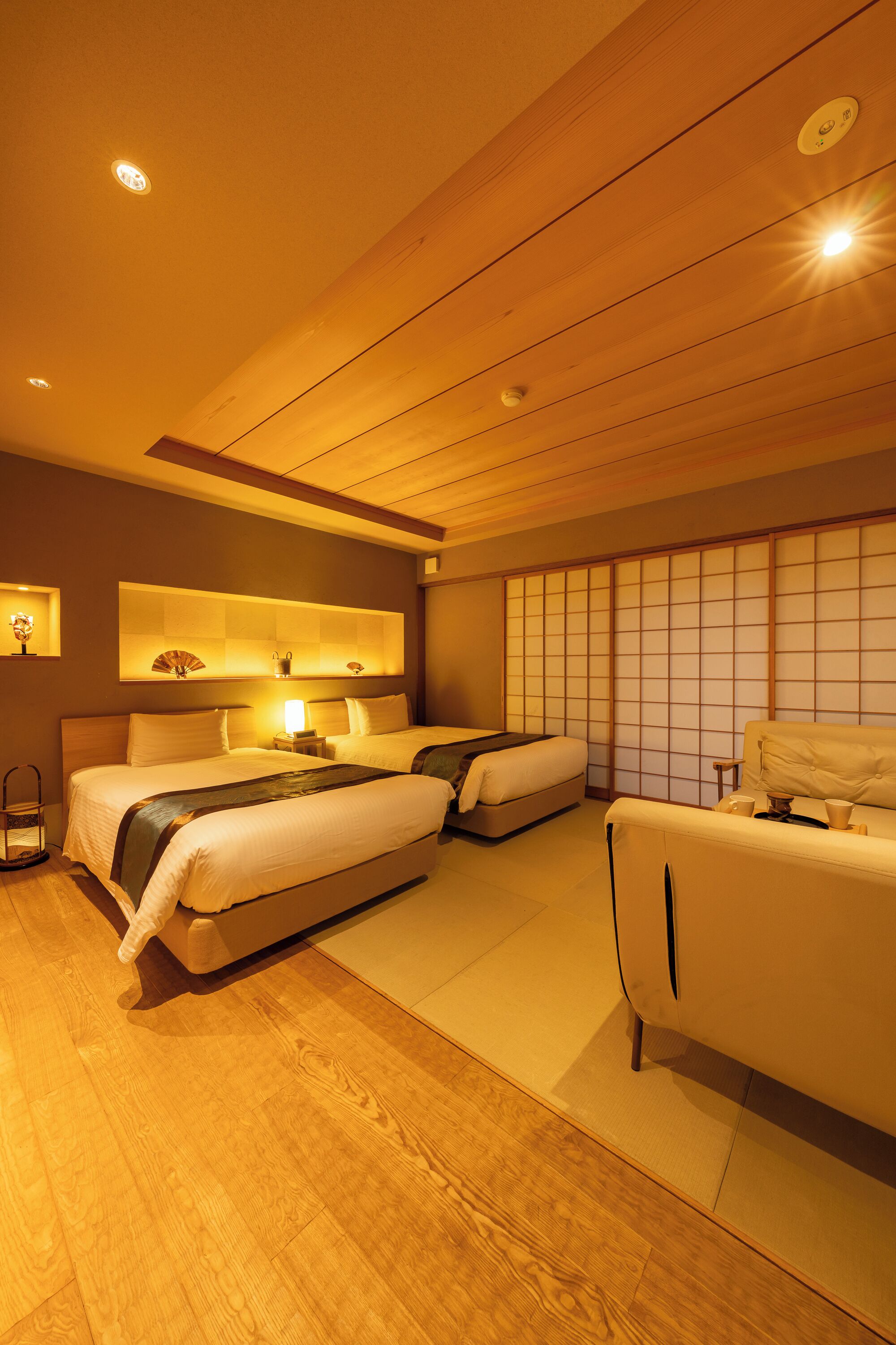 A hotel room with two beds, a sofa, a tatami mat floor, a shoji screen, and a ceiling fan.