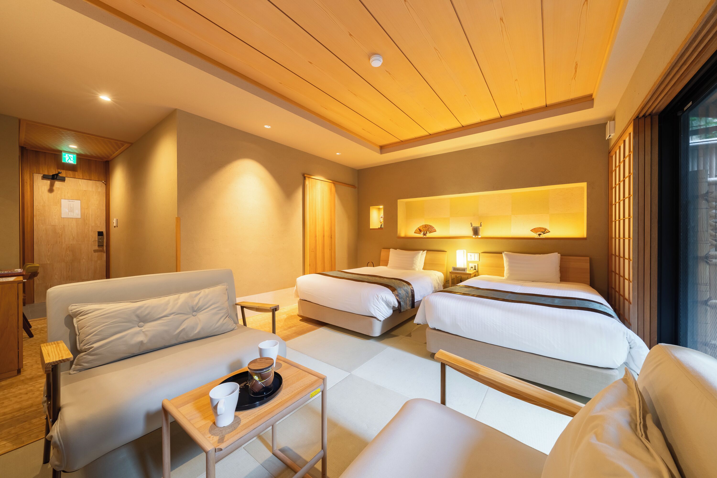 A hotel room with two beds, a sofa, a small table with a cup, and a window with blinds.