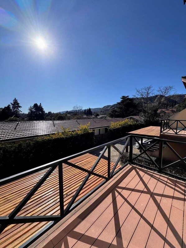 A wooden deck with a metal railing, overlooking a landscape with buildings and trees.
