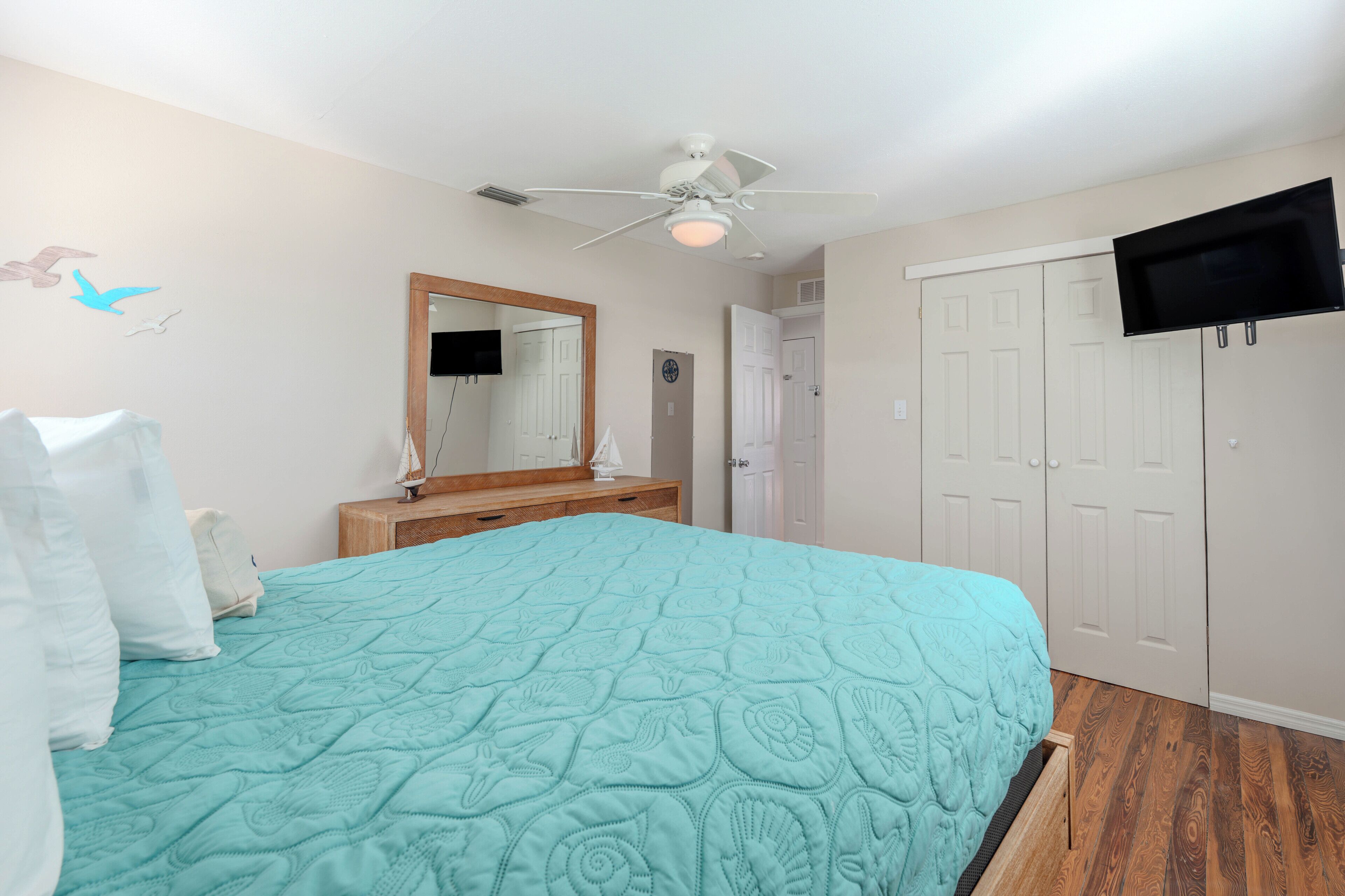A bedroom with a bed, a ceiling fan, a TV mounted on the wall, and a door.