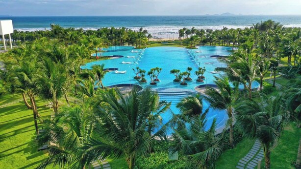 A large outdoor swimming pool with multiple sections, surrounded by palm trees and a view of the ocean. - THE ARENA CAM RANH (Bac Cam Ranh)