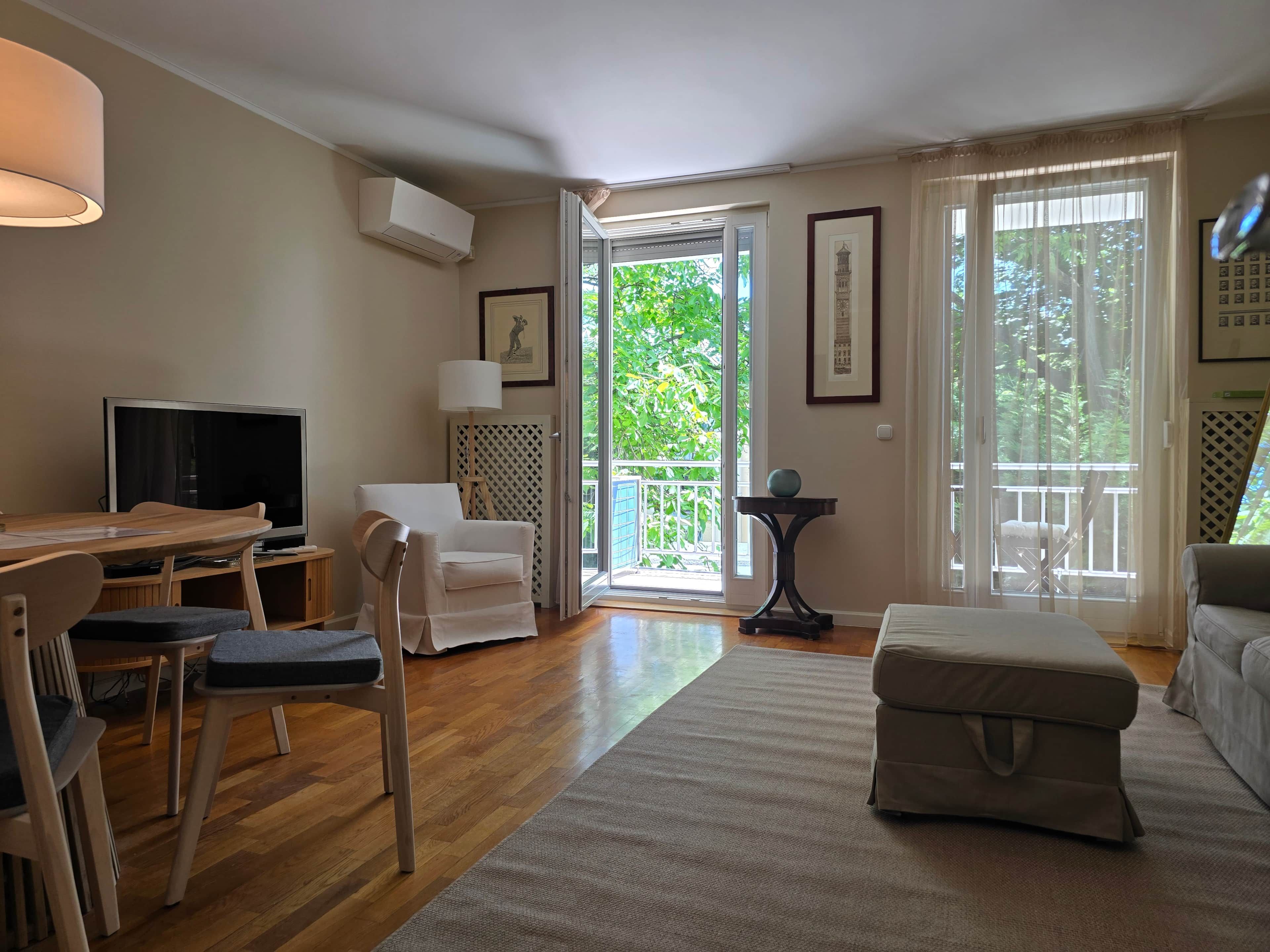 Una sala de estar con televisor, sofá, sillón, mesa y un ventanal con vistas a zonas verdes.