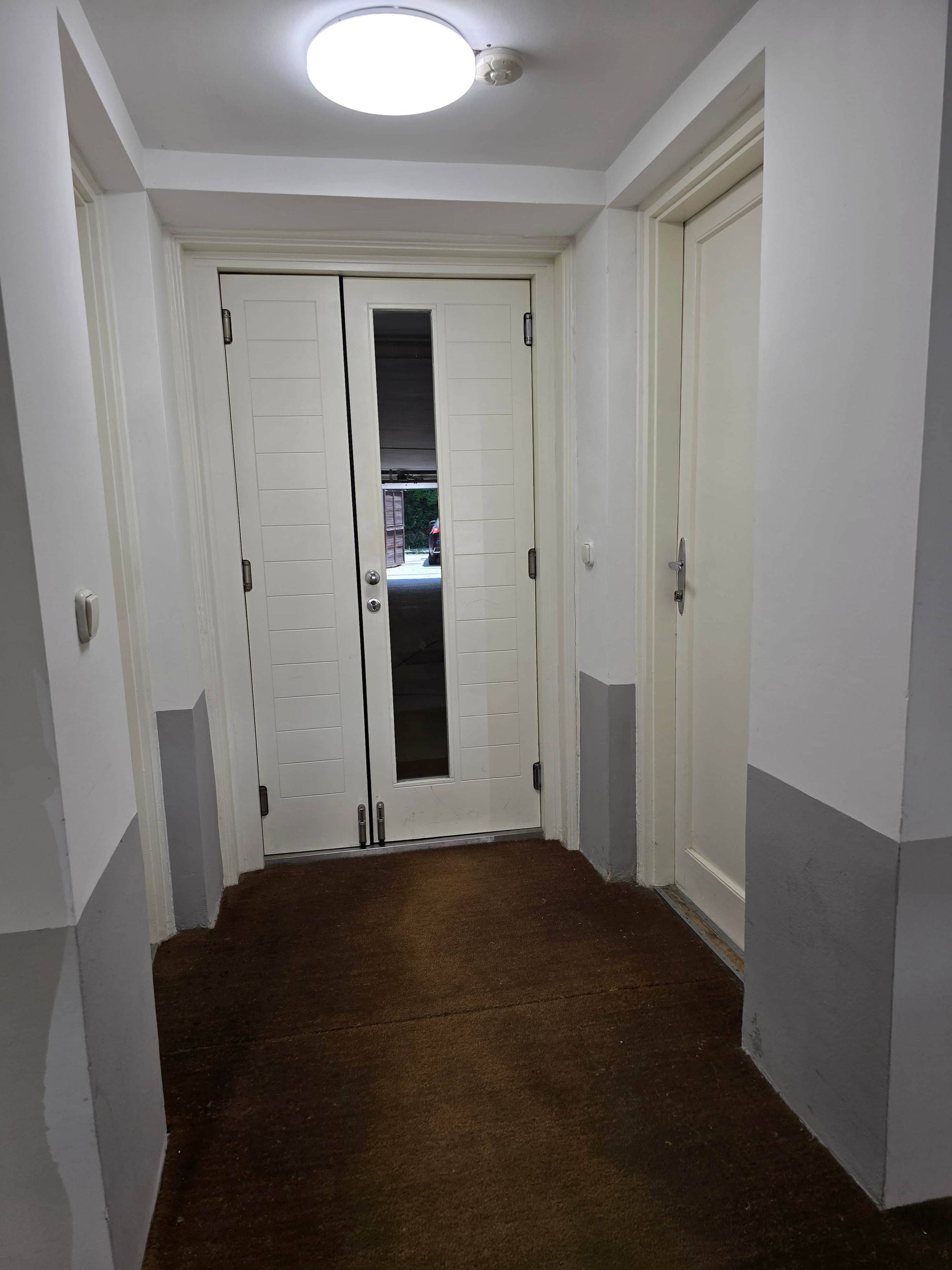 Un corridoio con due porte bianche, una delle quali con un vetro, e una luce a soffitto.