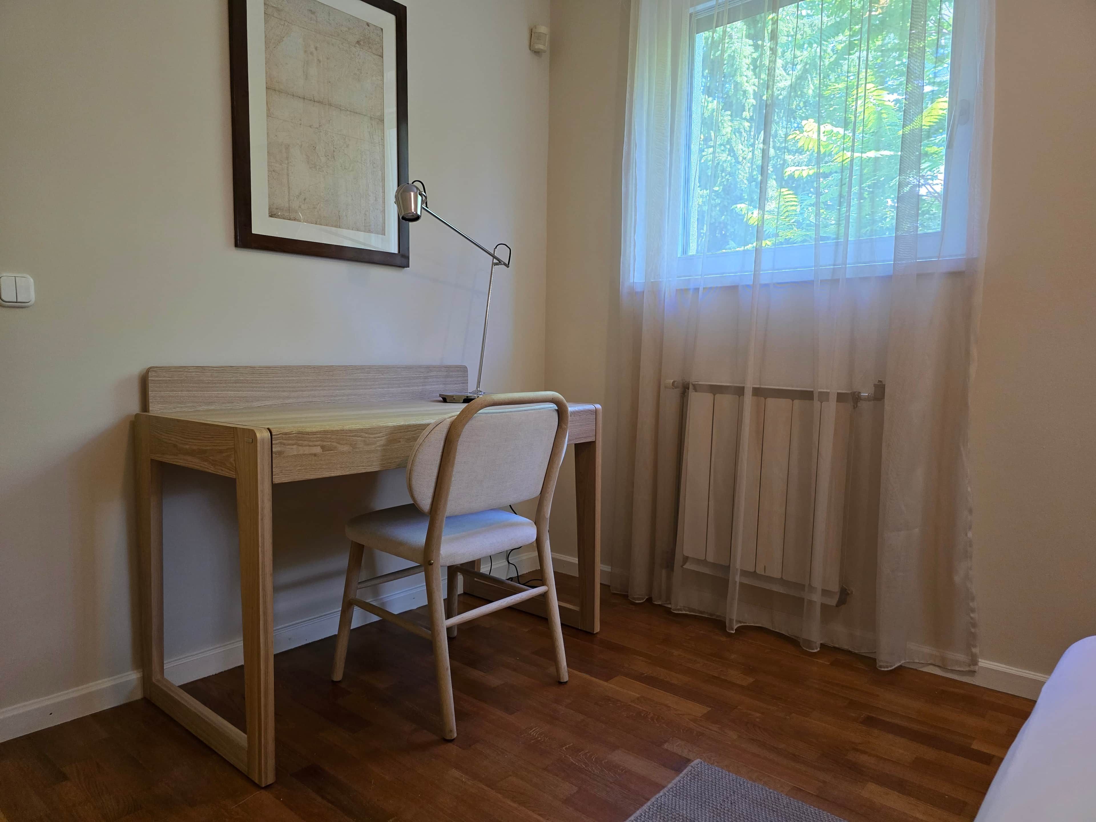 Une pièce avec un bureau en bois, une chaise, un radiateur et une fenêtre ornée de rideaux légers.