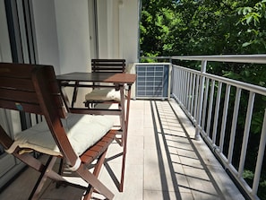 Una sedia e un tavolo in legno su un balcone con ringhiera bianca, una finestra e vegetazione sullo sfondo.