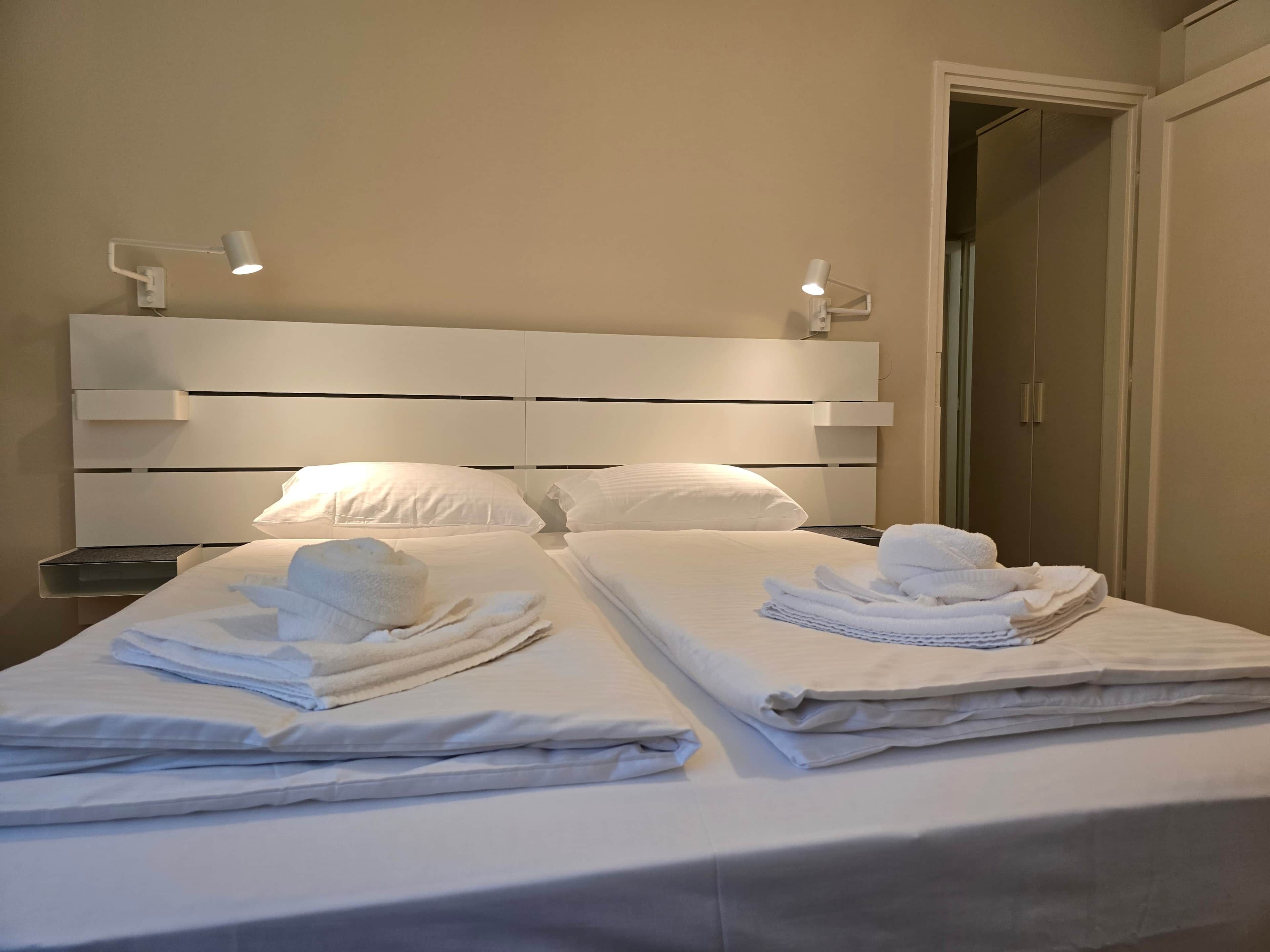 Quarto de hotel com duas camas, cada uma com toalhas dobradas e cabeceira com iluminação embutida.