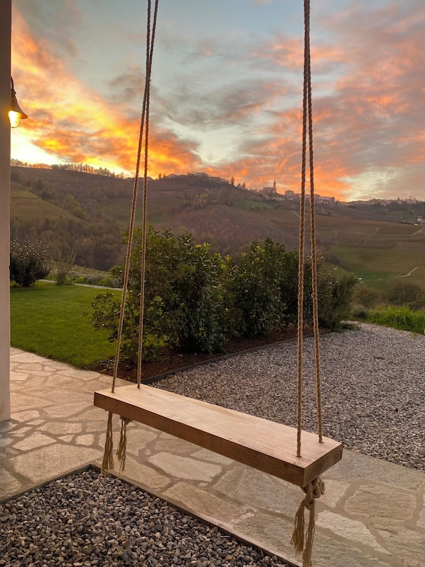 Une balançoire en bois installée sur une terrasse en pierre, avec vue sur un paysage vallonné et un coucher de soleil.