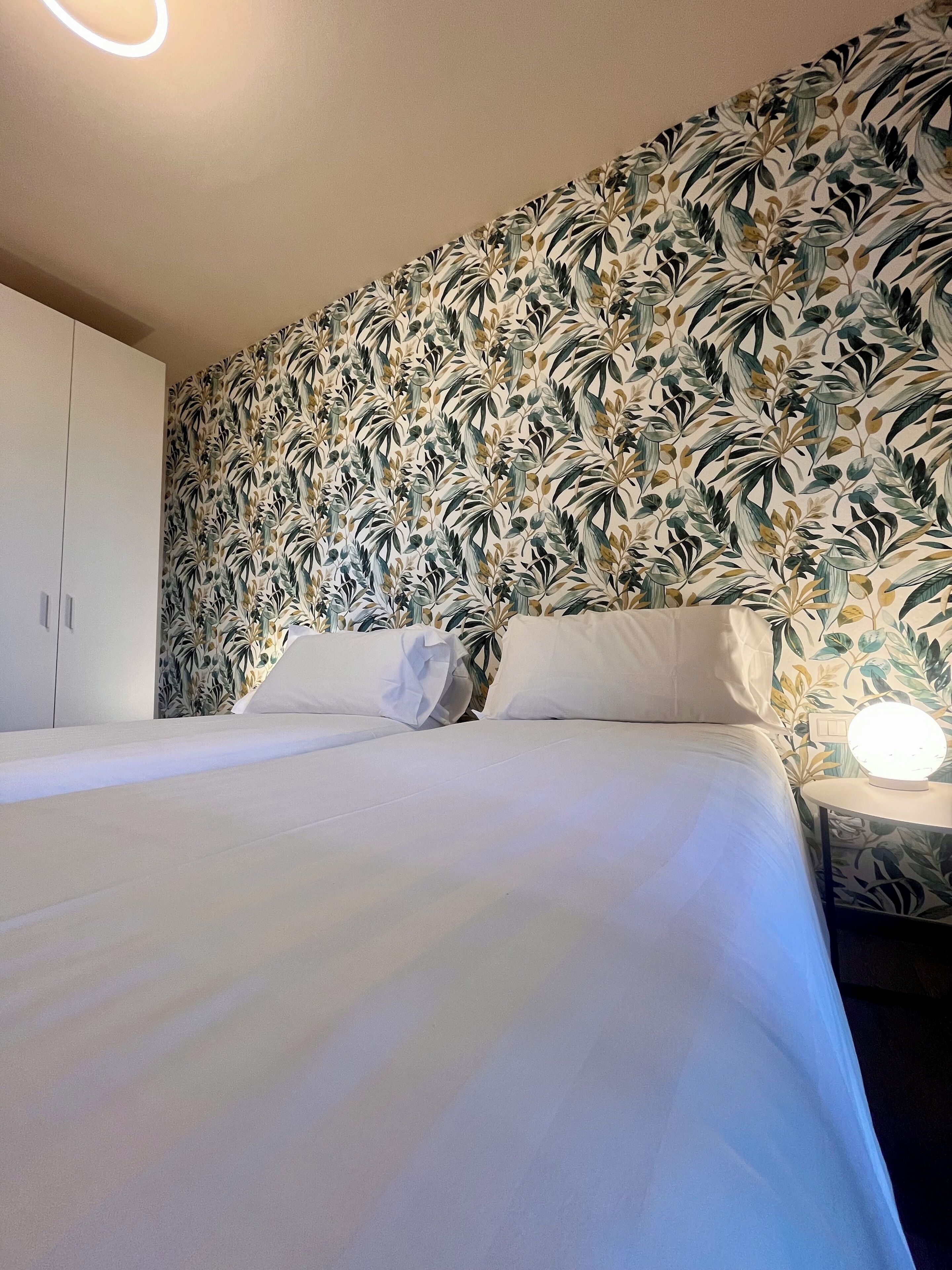 A bedroom with a floral wallpaper, a bed with white bedding, a bedside table with a lamp, and a wardrobe.