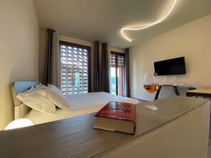 Habitación de hotel con cama, mesita de noche con un libro y una copa de vino, televisión y ventana con cortinas.