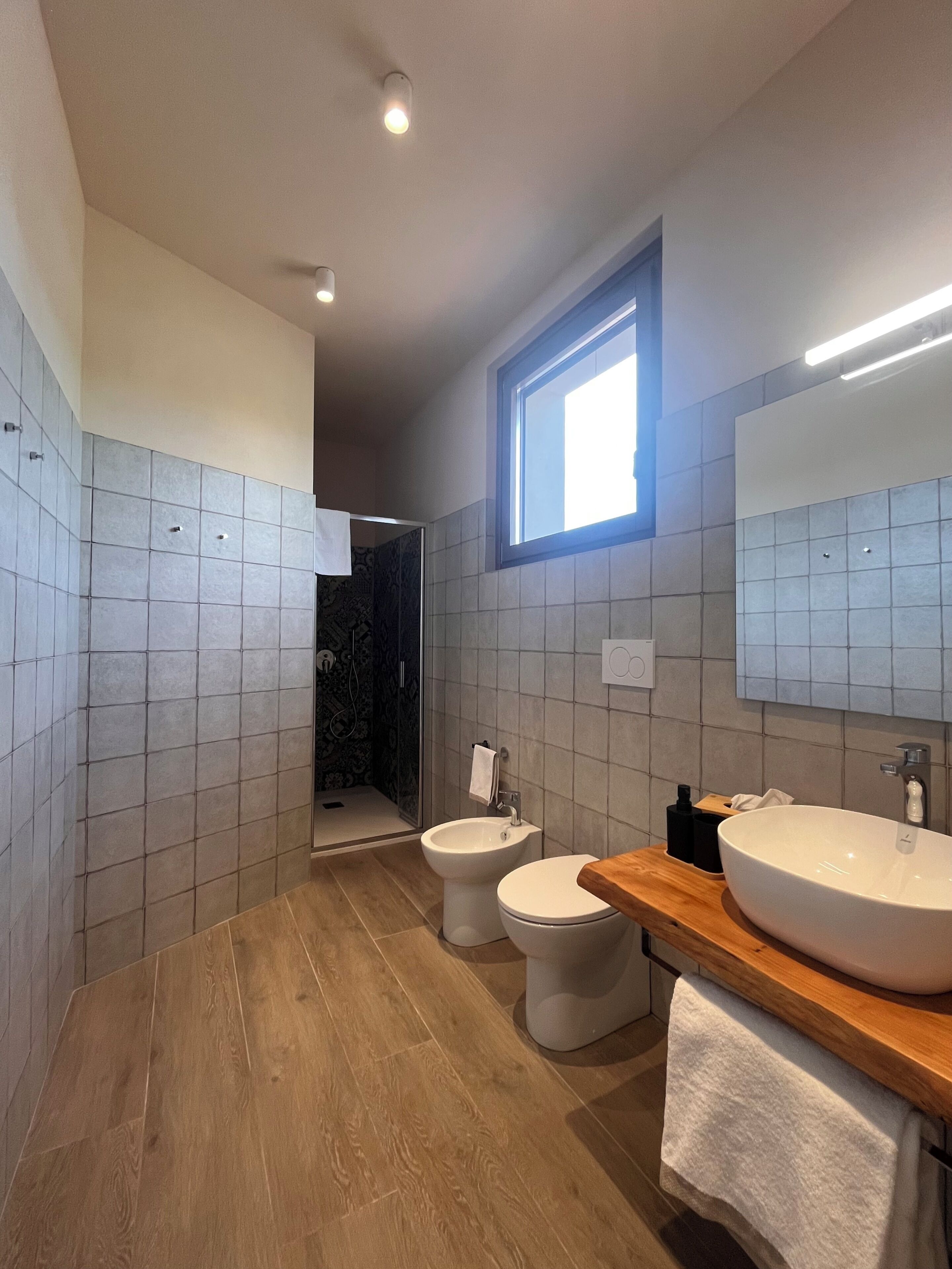 A modern bathroom with a wooden vanity, two toilets, a bidet, a shower area, and a window providing natural light.