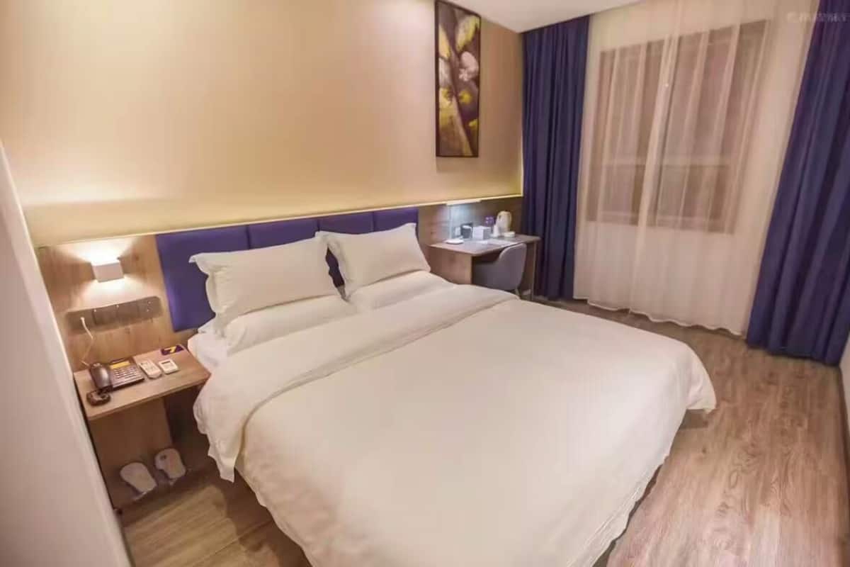 Quarto de hotel com cama, mesa de cabeceira, luminária e janela com cortinas.