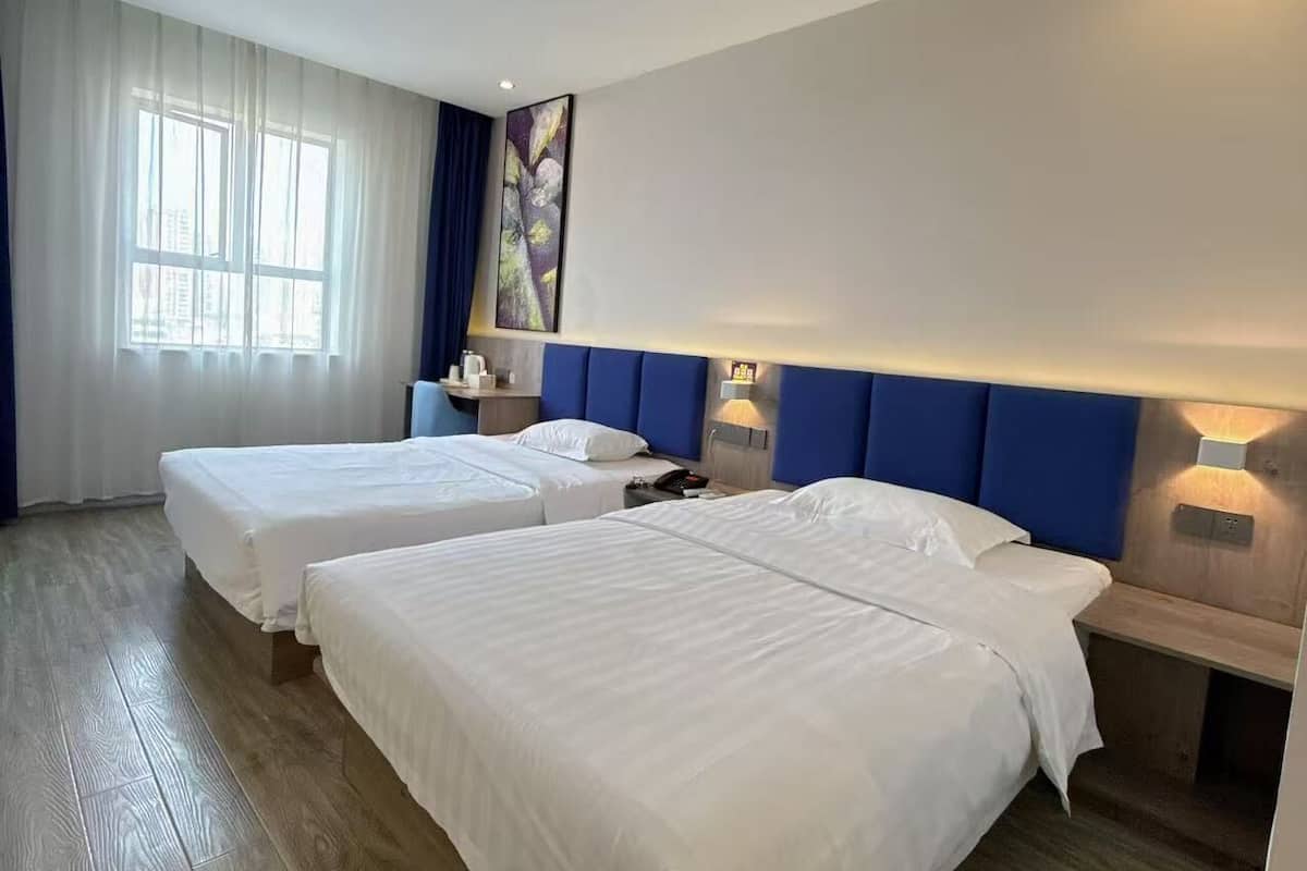 Quarto de hotel com duas camas, uma janela grande com cortinas, um quadro na parede e chão de madeira.