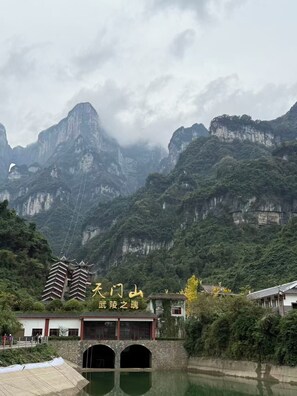 A traditional Chinese building with a stone bridge over a river, surrounded by lush greenery and mountains. - QīTiānYōupǐnZhāngjiājièDàyōngGǔchéngDiàn (Zhangjiajie)