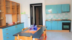 A compact kitchen with blue cabinets, a refrigerator, a stove, and a dining table with chairs. - Quadrilocale Angela N°6 - Salento Reservation (Torre Suda)
