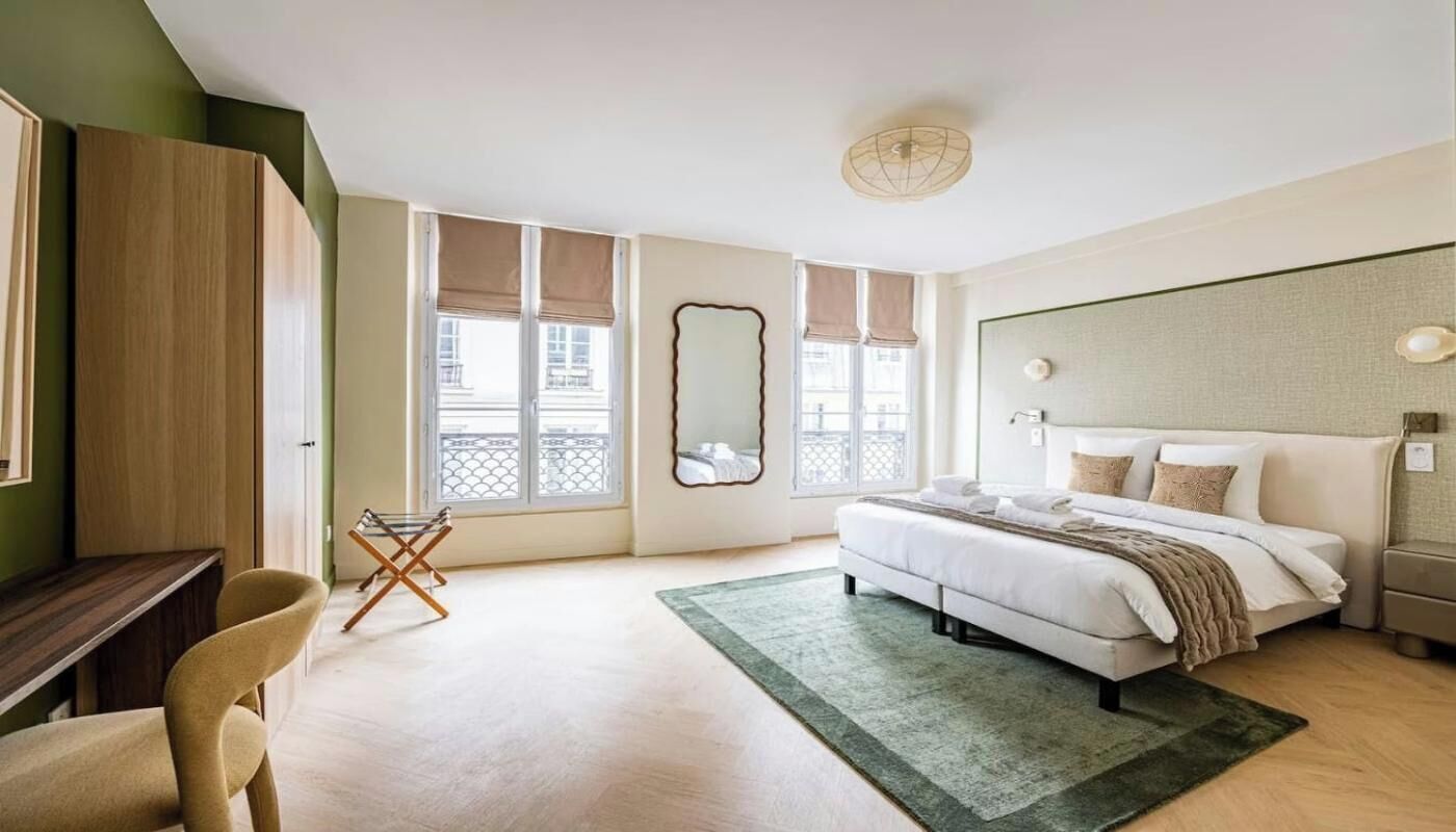 A bedroom with a bed, a desk, a chair, a window with a view, and a mirror on the wall.