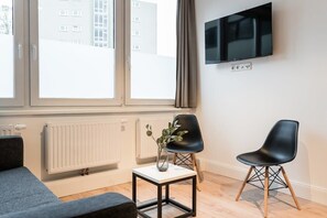 Ein modernes Zimmer mit einer Couch, einem kleinen Tisch mit einer Vase, einem schwarzen Sessel und einem an der Wand montierten Fernseher.
