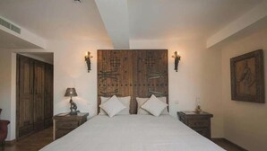 A bedroom with a large bed, wooden headboard, two bedside tables, a nightstand, a lamp, and a painting on the wall.