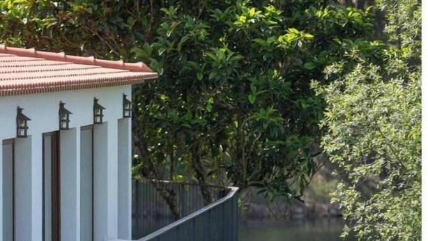A house with a balcony overlooking a body of water, surrounded by greenery.