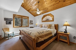 Un dormitorio con cama de madera, ventilador de techo, ventana con vistas a árboles, una silla y una mesita de noche con lámpara.