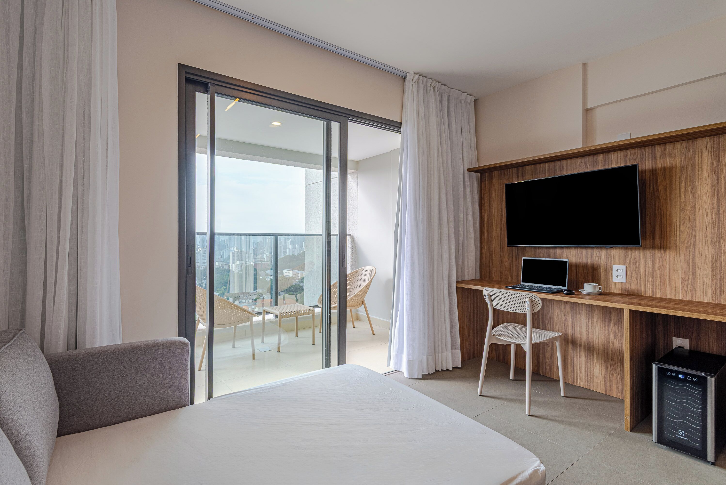 Une chambre d’hôtel avec un lit, un bureau avec un ordinateur portable, une télévision et une vue sur la ville, visible à travers une porte coulissante.
