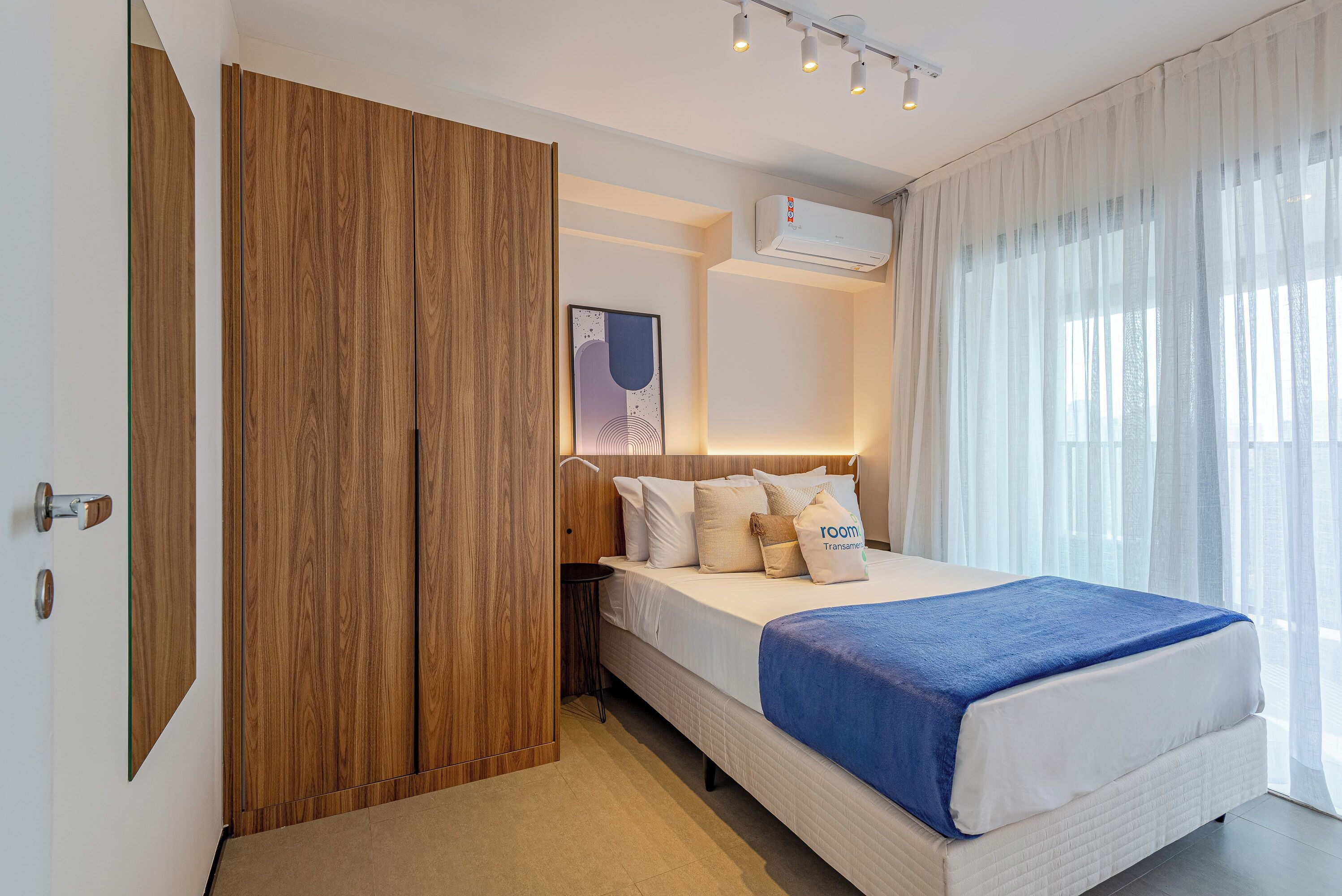 A bedroom with a bed, a wooden wardrobe, a bedside table, a wall-mounted air conditioner, and sheer curtains.