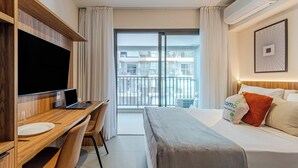 Ein Hotelzimmer mit Bett, Schreibtisch mit Laptop, Fernseher und Balkon mit Blick auf Gebäude.