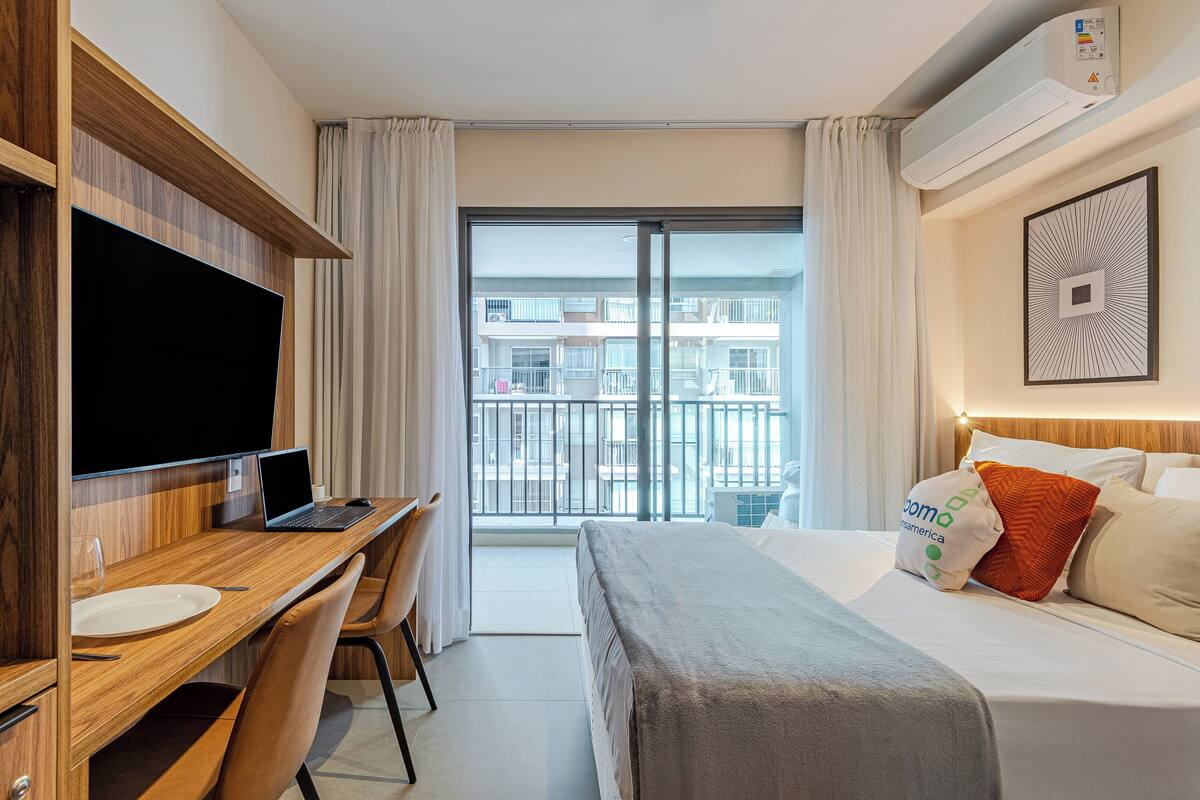 A hotel room with a bed, a desk with a laptop, a TV, and a balcony with a view of buildings.
