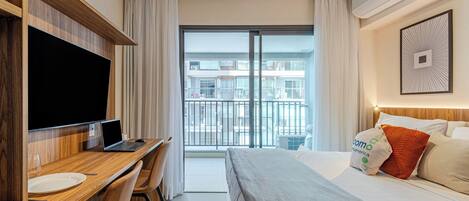 A hotel room with a bed, a desk with a laptop, a TV, and a balcony with a view of buildings.