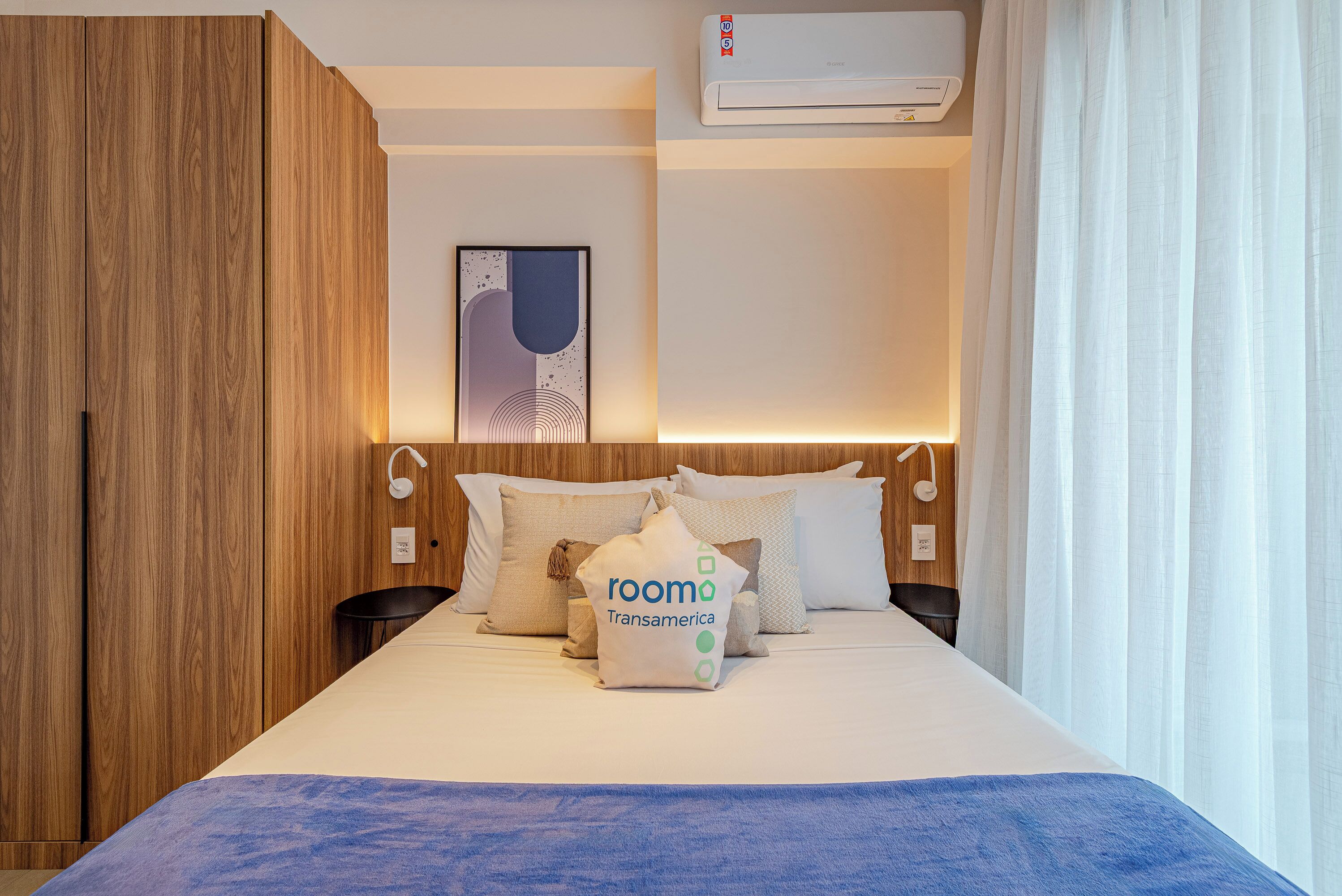 Una camera d'albergo con un letto, cuscini, un condizionatore d'aria a muro e un quadro incorniciato.
