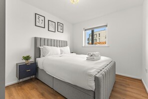A bedroom with a bed, bedside table, and a window with a view of buildings. - 6th Floor Balcony Flat Walk To Coop Arena & Etihad - Pass the Keys (Manchester)