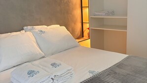 En pænt redt seng med hvide sengetøj og et gråt tæppe, en træsenggavl med hjerteformede dekorationer, og en indbygget garderobe med hylder.