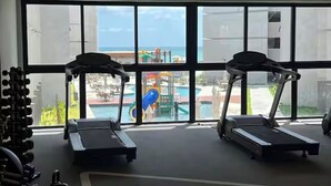 Et fitnesscenter med løbebånd og træningsudstyr, store vinduer med udsigt til en legeplads og en pool.