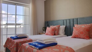 Quarto de hotel com duas camas, cada uma com colcha estampada, uma mesa de cabeceira com luminária e vista do céu por grandes janelas.