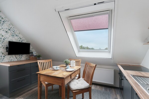Un coin repas avec une table en bois, des chaises, un téléviseur et une fenêtre avec un store rose.