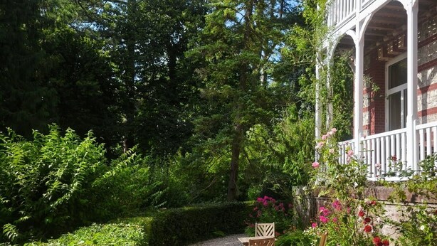 A gravel path leading to a wooden table and chairs set in a garden with dense foliage and a building with a balcony. - Villa Eliane (Étretat)