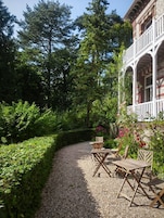 A gravel path leading to a wooden table and chairs set in a garden with dense foliage and a building with a balcony.