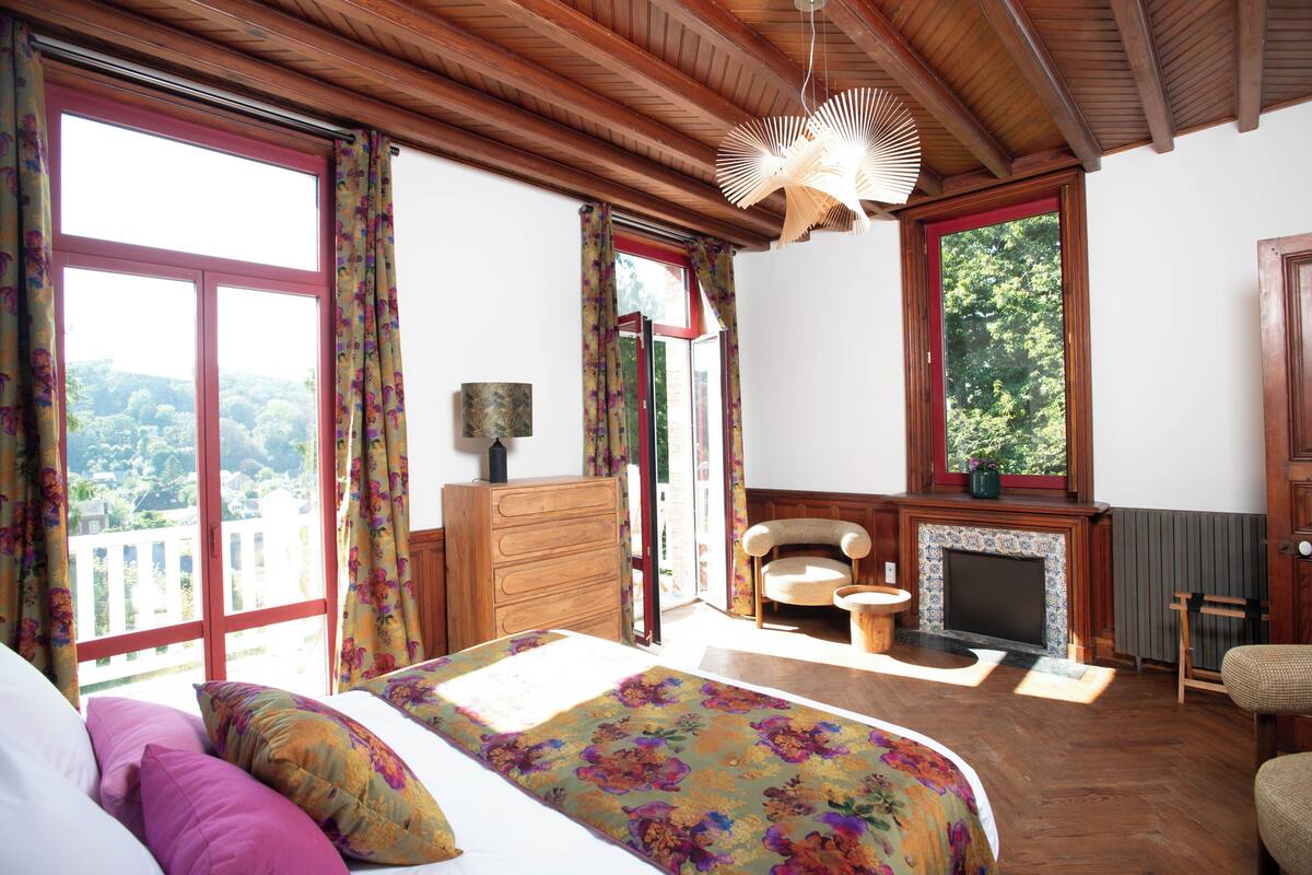 Un dormitorio con cama, cómoda, silla, chimenea y ventanas amplias.